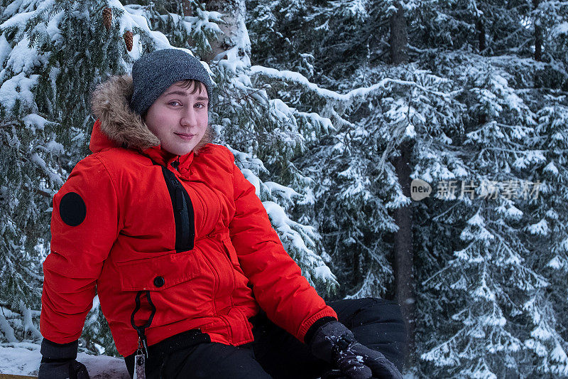 在白雪皑皑的森林背景下，一名年轻的白人男子穿着冬天的红色夹克，看着相机。