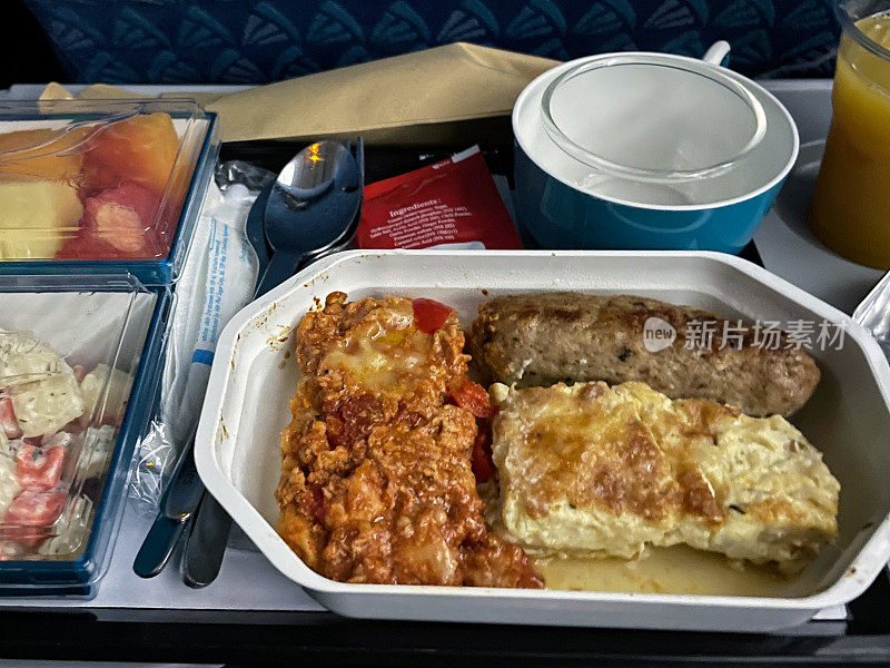 飞机食物托盘的全帧图像，包含早餐煎蛋卷，香肠，番茄和洋葱哈希，碗土豆和水果沙拉，塑料杯橙汁，咖啡杯，金属勺子和叉子，高视图