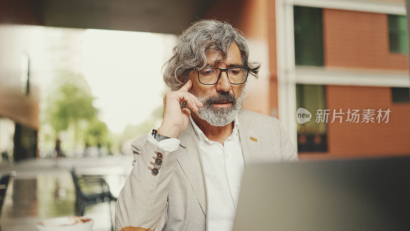 一个留着胡子、戴着眼镜、穿着灰色夹克的成熟商人坐在咖啡馆里。中年经理成功男士坐在户外的咖啡馆里，对着笔记本电脑