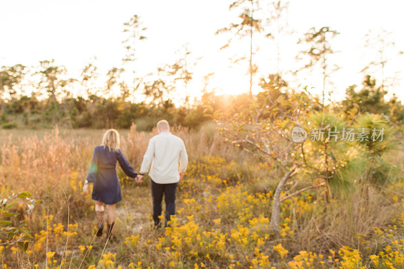 一个快乐的年轻的美国夫妇短金发和婴儿蓝眼睛穿着牛仔布和棕色皮革牛仔靴，散步和享受时间一起在田野的黄色野花和潘帕斯草在黄金时刻在朱庇特农场，佛罗里达州