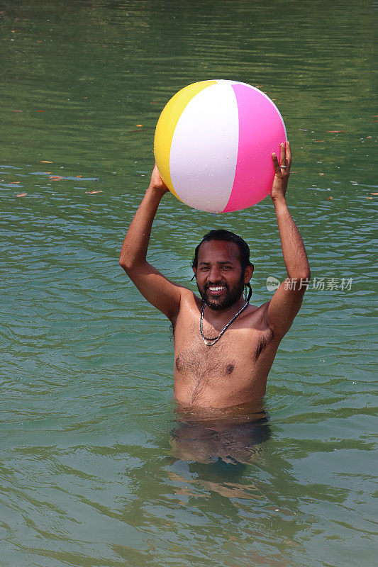 特写照片:一名印度男子穿着冲浪短裤站在齐腰深的泻湖平静的水中，头上举着彩色的塑料沙滩球，这是前景的焦点