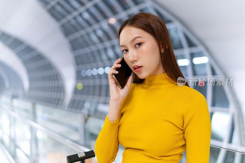 一个年轻的亚洲女人在机场候机厅打电话，看着镜头，她身边放着一个手提箱。出差的年轻亚洲女商人。年轻女子的生意在移动。随时随地的数字银行。