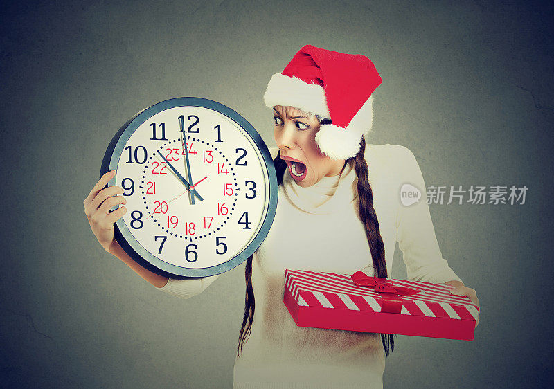 特写肖像着急紧张的年轻女子戴着红色圣诞老人帽拿着时钟礼盒