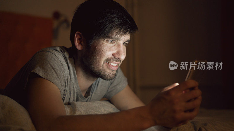 年轻微笑的男子在晚上躺在家里的床上用智能手机浏览社交媒体的特写