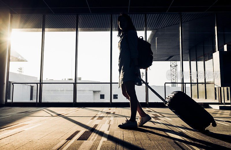 日出时分，年轻女孩带着行李走在机场航站楼窗口的剪影，旅行理念，旅行生活方式
