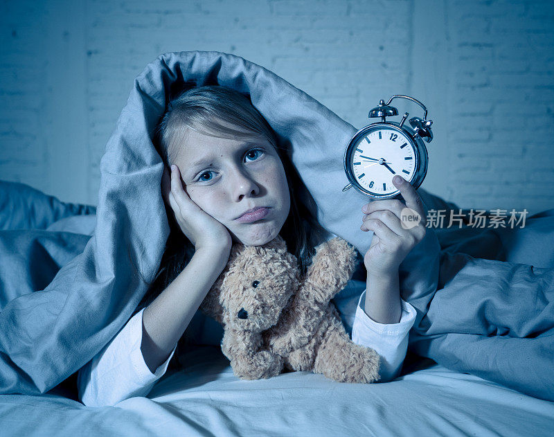 可爱的失眠小女孩躺在床上，闹钟看起来很累，有睡眠问题，晚上睡不着觉，或在儿童失眠、焦虑和睡眠障碍中过早醒来。