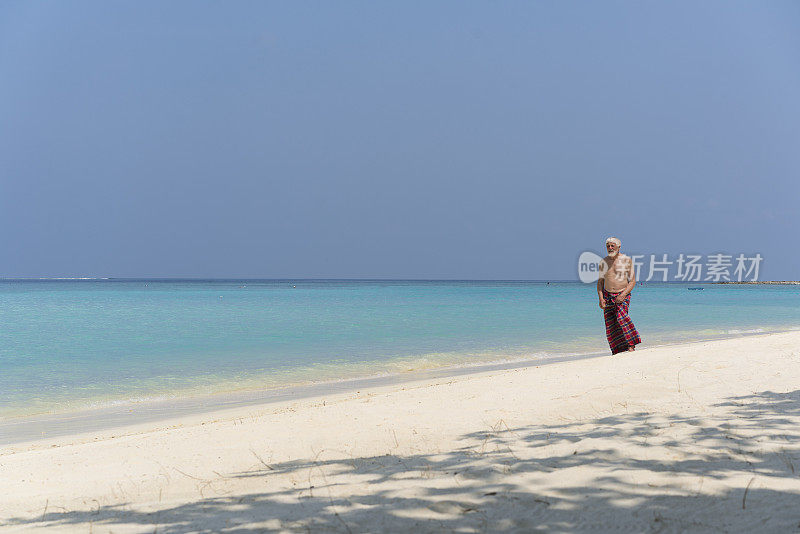 游客，身着纱笼的老年男子，走在马尔代夫Gulhi岛的“比基尼海滩”上
