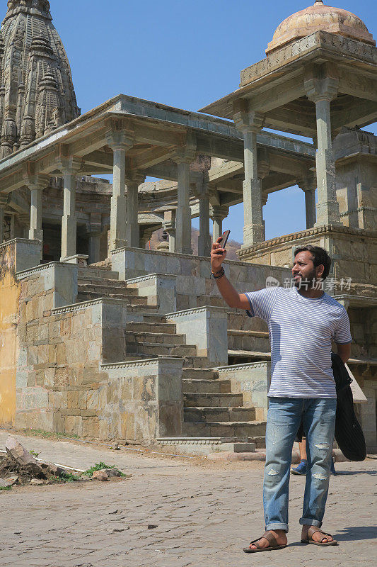 印度游客度假印度，男子自拍用手机相机，英俊的印度印度男子模特酷时尚发型观光寺庙，破洞牛仔裤凉鞋自拍照片社交媒体发帖，自拍度假