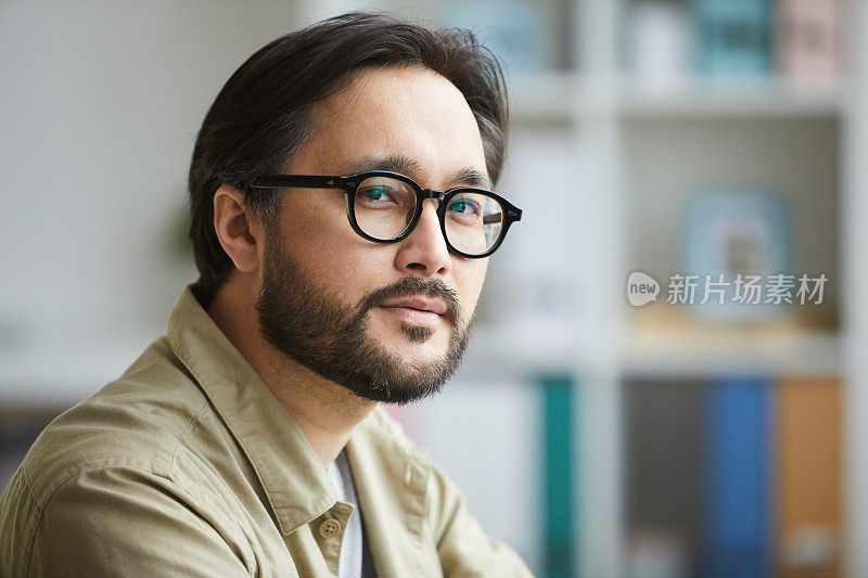 一幅严肃自信的年轻亚洲男子的肖像，留着胡子，戴着眼镜，穿着衬衫在办公室工作