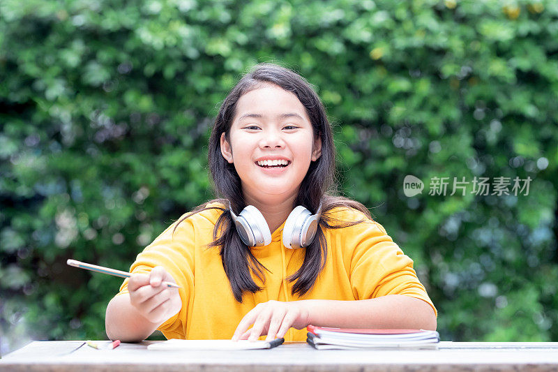微笑的亚洲小女生在公园里做作业，她的脖子上戴着耳机