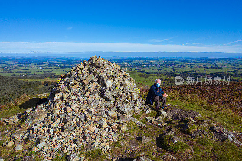 高角度的观点，一个活跃的老年人戴着口罩，而坐在旁边的一个石碑上的苏格兰山顶