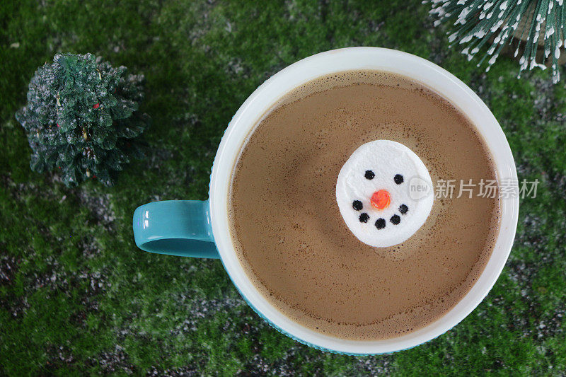 近距离拍摄的棉花糖雪人的脸漂浮在热腾腾的巧克力杯中，背景是圣诞树森林效果，居高临下的视野