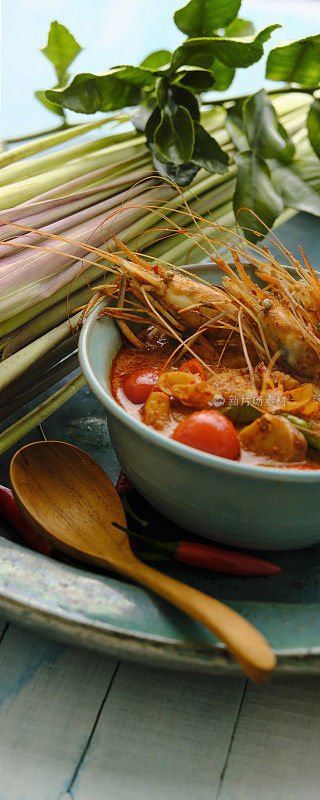 在国际上享有盛誉的泰国辣椰汤“冬阴功”，在蓝色的碗里与汤勺形成鲜明对比，柠檬草、青柠叶和红辣椒放在蓝色的陶瓷盘子里，在蓝色的木制桌子的背景上。