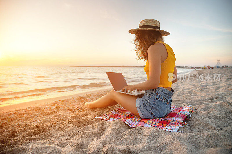 日落时分，一位身材苗条、身穿牛仔短裤、黄色背心、头戴草帽的年轻女士坐在沙滩上的毯子上，用笔记本电脑工作。