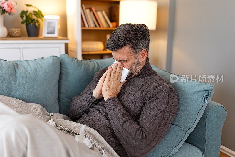 生病的悲伤的男人坐在家里的沙发上流鼻涕流感冠状病毒大流行covid流行病打喷嚏。不舒服的家伙感觉不好发烧病毒病症状室内