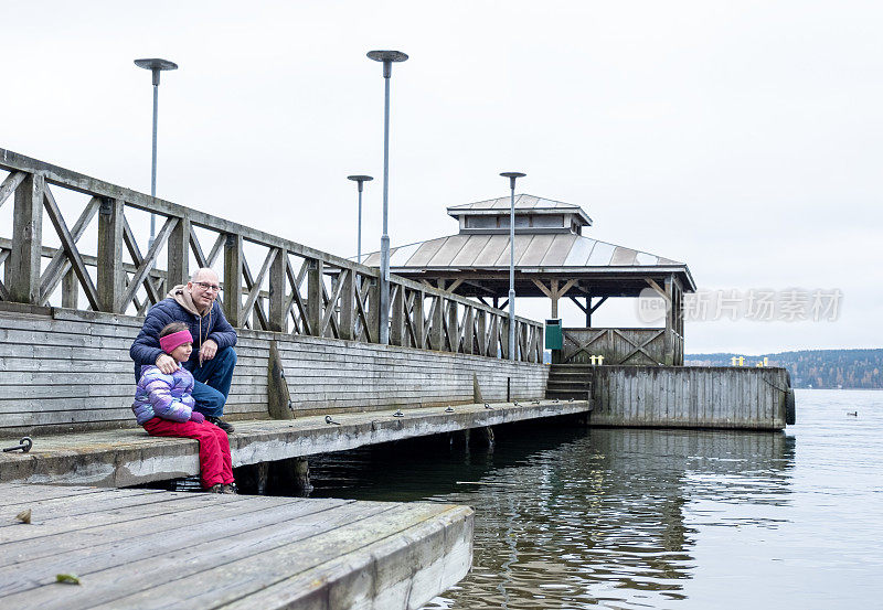 一个7岁的白人女孩和一个中年男子坐在湖边的木码头上做梦。秋天的心情。