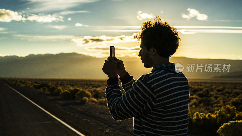 一个年轻人，旅行潮人，在内华达州的沙漠道路上用智能手机拍摄日落