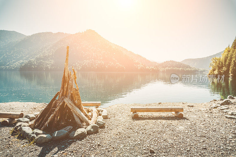 准备点燃一个巨大的篝火和长凳上的原木在一个美丽的湖岸边，清澈的水被山和森林包围在明亮的夏季太阳的背景