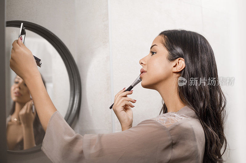 美丽的年轻女性看着镜子化妆。在浴室里