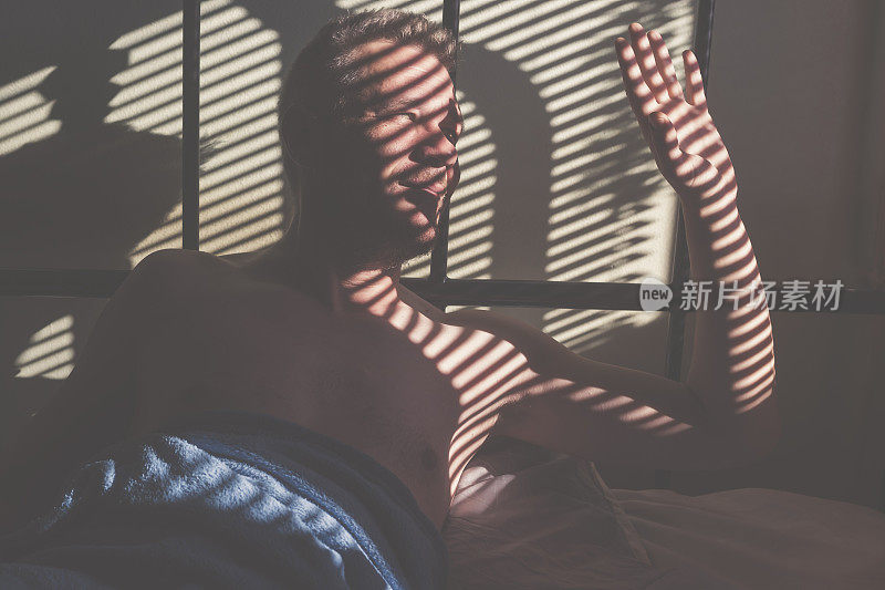 英俊的成年男子早晨醒来，阳光透过百叶窗照亮他的阳光