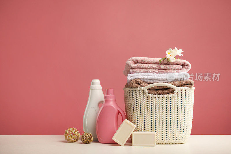 粉红色背景上有毛巾、小苍兰花、洗涤剂、竹球和肥皂的洗衣篮
