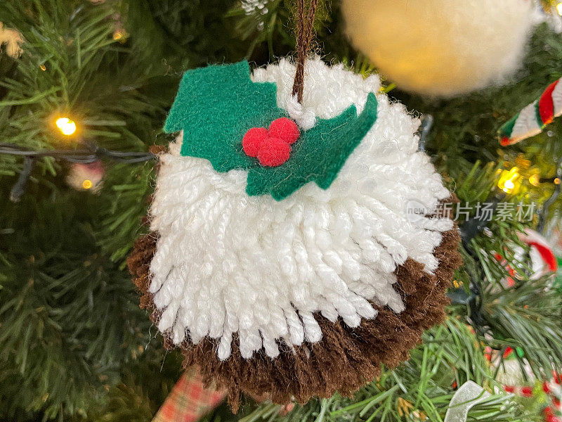 特写图像精心制作的羊毛圣诞布丁绒球在人造圣诞树树枝，绿色云杉针，模糊的白色精灵灯背景