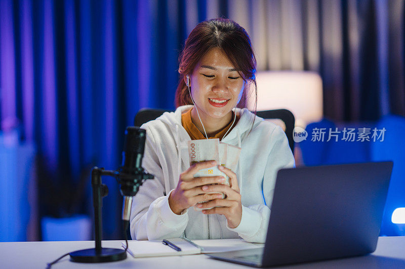 年轻的亚洲妇女拿着钱银行票据和录制播客在她的笔记本电脑在家庭工作室在晚上。