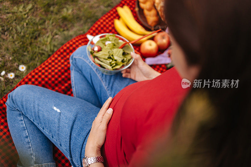 年轻漂亮的孕妇坐在红色的毯子上，吃着混合蔬菜沙拉。