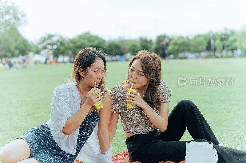 两个年轻的亚洲女性朋友在公园里喝着饮料，吃着野餐，看着对方