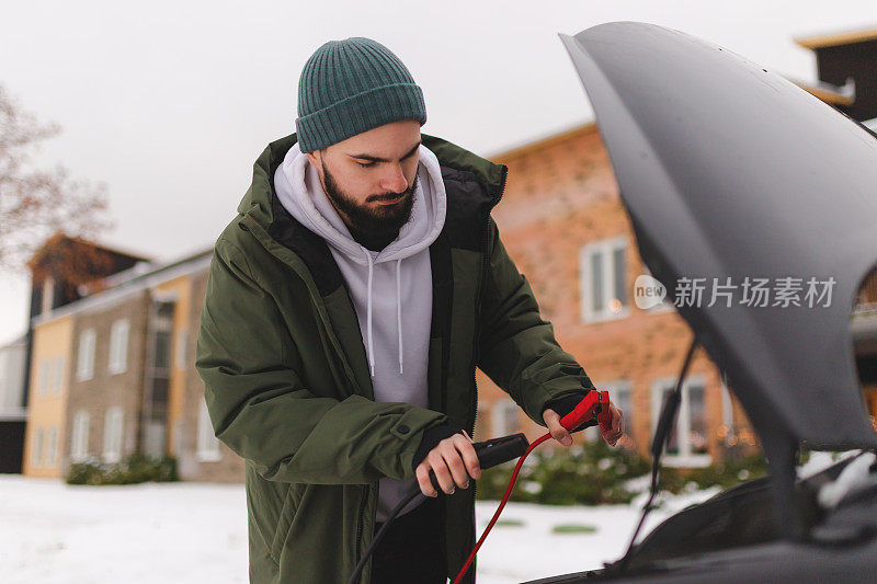 穿着冬装的年轻人在冬天用电池电缆启动他的汽车
