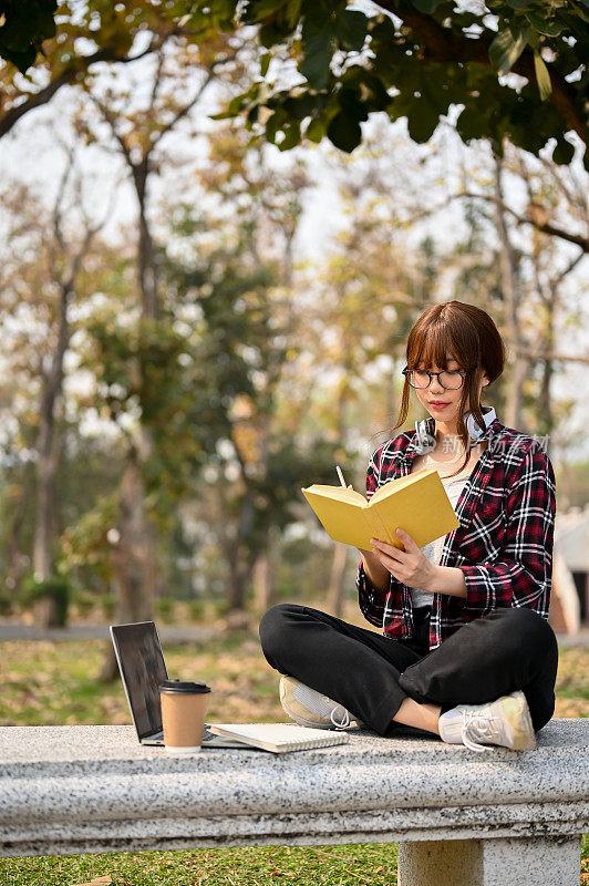 年轻迷人的亚裔女大学生在校园公园的长椅上看书