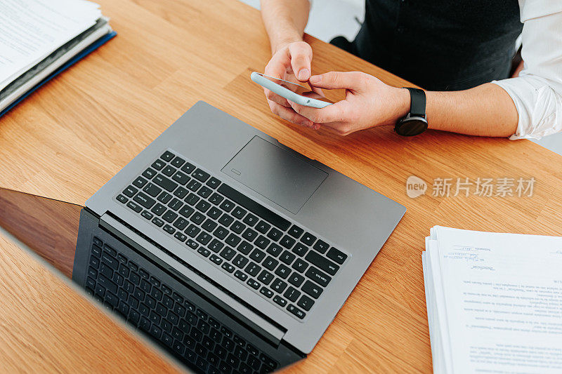 一名无法辨认的男子手持智能手机，在光滑的黑色笔记本电脑键盘上打字