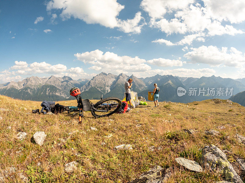 一对夫妇在瑞士阿尔卑斯山骑自行车，日落时分在山顶搭起帐篷