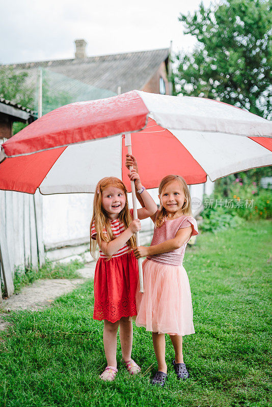 快乐有趣的孩子们在夏天的阵雨或大雨中在后院拿着大沙滩伞。女孩们穿着连衣裙在春天的公园里散步，享受着雨水。孩子们在玩耍，接雨滴。