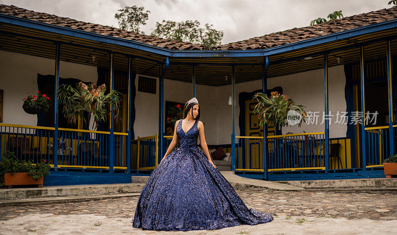 身着quinceañera连衣裙的年轻女子站在一栋典型的哥伦比亚房子中间，头戴皇冠，望向一旁