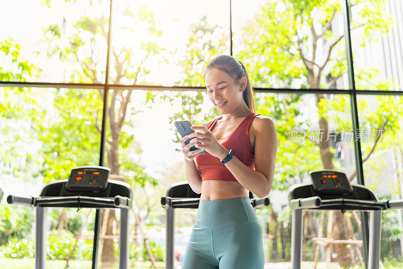 亚洲女性在阳光灿烂的室内健身俱乐部用智能手机休息，拥有花园景观