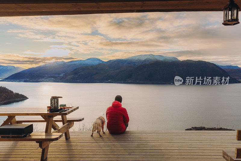 在挪威的日落时分，一名男子带着狗在露台上凝视着冬季峡湾的美景