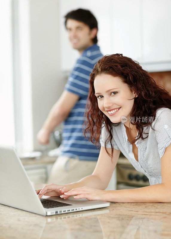 年轻女性在厨房拿着笔记本电脑，男人在后面