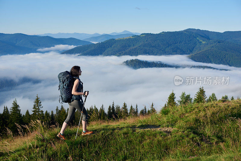 后视图的运动女孩徒步旅行者背包和徒步旅行的棍棒远足在乡村小径上的一个小山，看着远处，享受早晨的阴霾在山谷，森林在模糊的背景