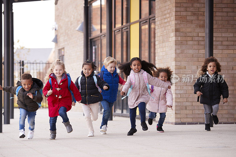 一个快乐的多民族的年轻学校的孩子们穿着大衣，背着书包和他们的同学在他们的幼儿学校大楼外面的人行道上跑步