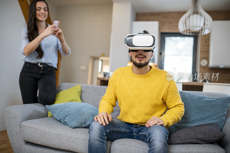 一名年轻人用虚拟现实头盔和女友用智能手机拍摄他的视频