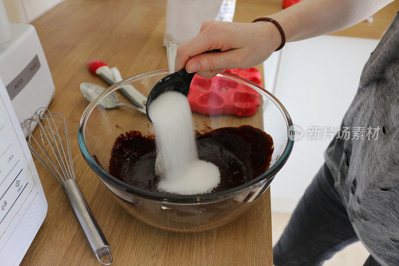 这是一名白人少女将白砂糖配料倒入玻璃搅拌碗中，搅拌碗中含有黄油、面粉和可可的巧克力蛋糕面糊