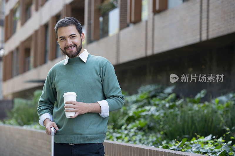 一个拉丁裔的年轻人走在街上，穿着一件绿色毛衣，一只手拿着咖啡，另一只手拿着旅行箱。似乎微笑着看着镜头