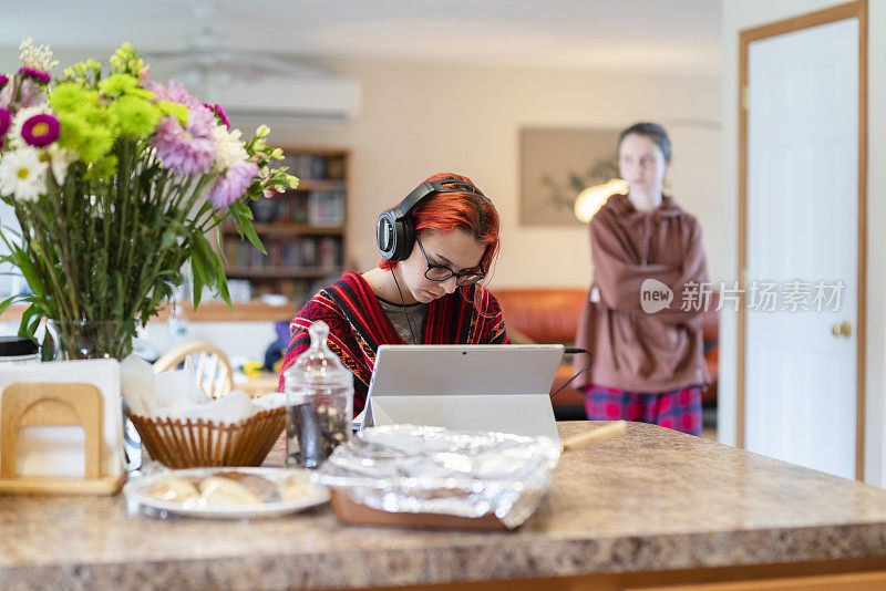 在家学习并不总是舒适的。这个十几岁的女孩戴着耳机在远处上课，在厨房柜台边用笔记本电脑工作，这时她的大姐正从后面走进客厅。