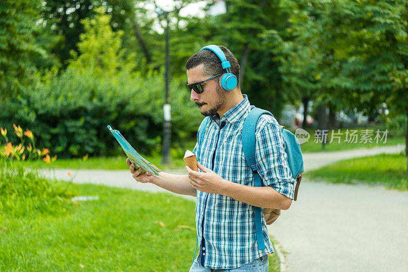 一个穿着格子衬衫的帅哥正在城市公园里散步，一边听着音乐，一边吃着冰淇淋提神，一边看地图。