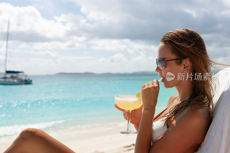 一个女人在热带海滩的柚木躺椅上喝着鸡尾酒