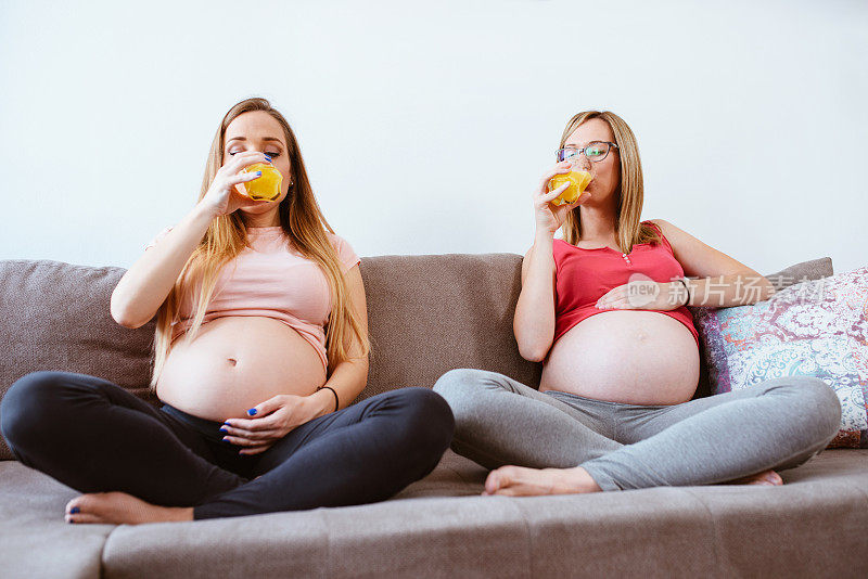 两个开心的孕妇坐在沙发上喝着新鲜的橙汁