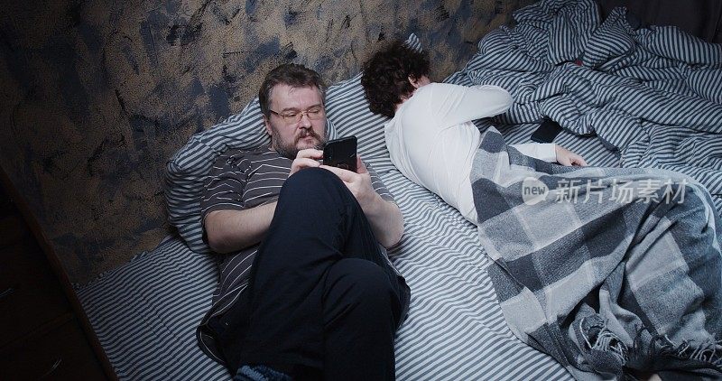 成熟的妻子试图睡觉，而丈夫在床上使用她的智能手机