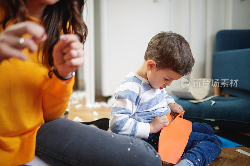 蹒跚学步的孩子坐在他妈妈旁边，在地板上用剪刀剪纸