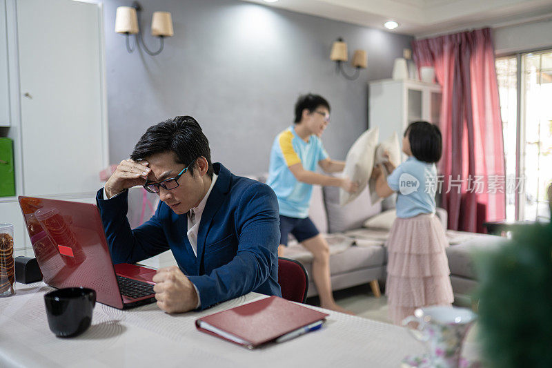 亚洲华人在家工作。孩子们在玩枕头大战。父亲失去了注意力和沮丧。